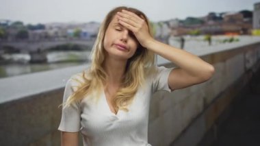 Şehirdeki bir nehrin kenarında duygularını ifade eden bir kadın. Sarı saçlı, kederli bir ifade. Şehir ortamında günlük beyaz gömlek giyiyor..