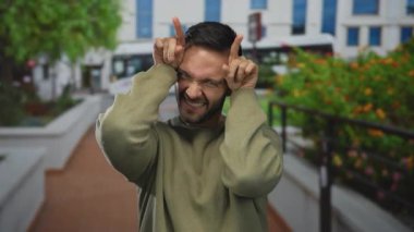 İspanyol bir adam eğlenceli boynuzlar jest yapıyor parmaklarıyla şehir merkezinde, neşeli bir ifade ve gençlik enerjisi sergiliyor..