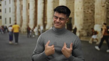 Gözlüklü genç adam hareketli bir caddede gülümsüyor ve el kol hareketi yapıyor. Arka planda antik taş mimarinin önünden geçen insanlar var..