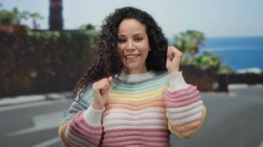 Sahilde güneşli bir günün tadını çıkaran bir kadın. Arka planda deniz var. Renkli bir kazak giyiyor..