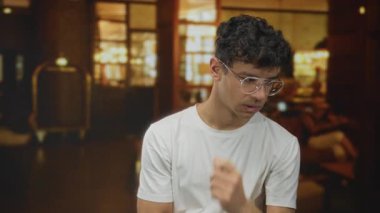 Beyaz gömlek giyen ve gözlüklü bir adam lobide sıcak ışıklarla çeneye dokunuyor..