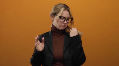 Kadın, izole edilmiş turuncu arka plana, gözlük takarak ve siyah spor ceket giyerek kafa karıştırıcı bir şekilde duygularını sergiliyor..