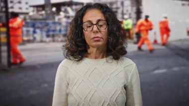 Kıvırcık saçlı yaşlı İspanyol kadın, arka planda turuncu üniformalı işçilerle bir inşaat sahasında üzgün görünüyor..