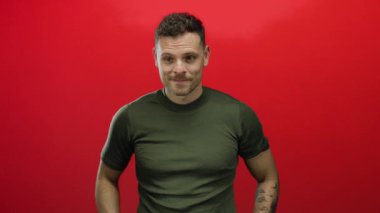 Genç İspanyol bir adam, izole edilmiş kırmızı bir arkaplan karşısında parmağını hareket ettiriyor, kolunda yeşil gömlekli, görünür bir dövmesi olan davetkar bir yüz ifadesi sergiliyor..