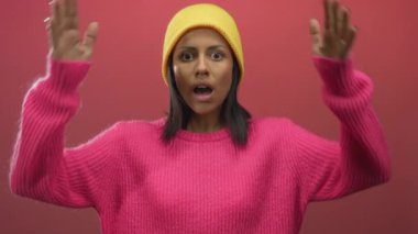 Parlak pembe bir süveter ve sarı bir bere giyen, cesur bir görsel ortamda duygu ve tarz sergileyen canlı kırmızı arka planda bir kadın..