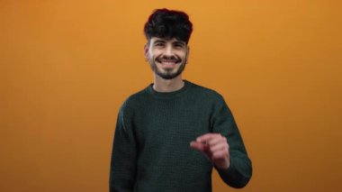 Turuncu arka planda örgü süveter giymiş gülümseyen genç adam küçük ya da küçük parmaklarını kaldırmış bir jest gösteriyor..