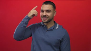Mavi polo tişört giyen, gülümseyen ve canlı kırmızı stüdyoda parmağıyla işaret eden İspanyol bir adam..