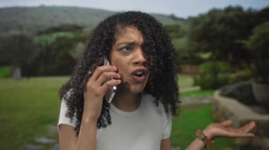 Beyaz gömlekli kadın parkta konuşurken elini kaldırarak ve ağzını açarak akıllı telefonu kulağına tutuyor..