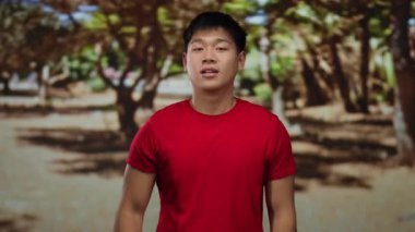 Kırmızı tişörtlü genç Çinli adam açık hava parkında iyi bir jest yapıyor..