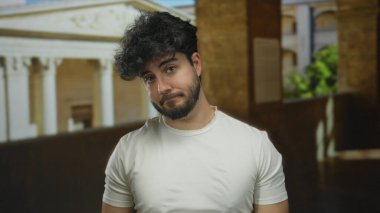 Sakallı genç adam, arka planda tarihi bir mimariyle eski bir üniversitede dikilirken şüpheli görünüyor..