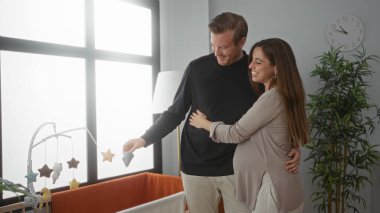 Hamile kadın ve erkek, sıcak bir yuva ortamındaki bebek beşiğine hayrandır. Duyguları ve aşkları ifadelerinde açıkça görülüyor..