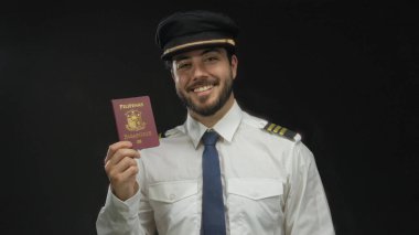 Siyah arka plan, seyahat ve profesyonel havacılık temalarına karşı Filipin pasaportu tutarken sakallı genç bir pilot gülümsüyor..
