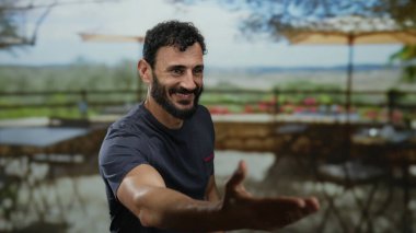 Lokantada baş parmağıyla gülümseyen bir adam açık havada bulanık masalarla arkadaşça pozitif yemek atmosferini yakalıyor..