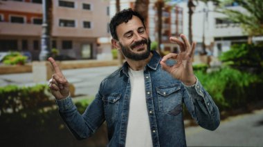 Sakallı İspanyol bir adam güzel hareketler yapıyor güneşli bir sokakta açık havada, modern binaların arka planına sahip kentsel bir çevrede mutluluk ve günlük moda sergiliyor..