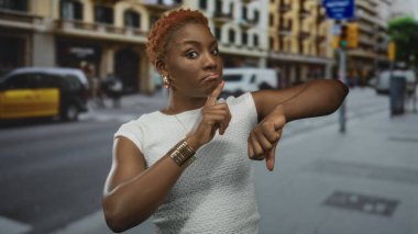 Beyaz uçlu bir kadın trafik ışığı ve mağazaların yakınındaki işlek bir caddede bileğini işaret ediyor..