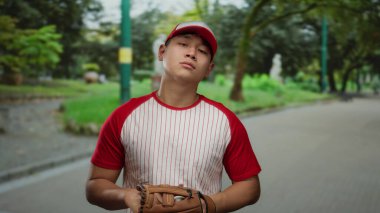 Beyzbol formalı, eldivenli ve toplu genç bir adam parkta duruyor, açık hava ortamının ortasında odaklanmış bir ifade sergiliyor, sporu doğa ve gençlikle harmanlıyor..