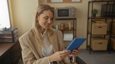 Kadın ofiste otururken selfie çekiyor. Arka planda raflı bej ceket ve mikrodalga fırın var. Günlük iş ortamını gösteriyor..