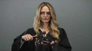 Siyah cüppeli, sarı saçlı kadın yargıç gri bir arkaplan üzerinde tokmak tutuyor, otoriteyi ve adaleti iletiyor..