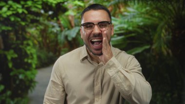 Gözlüklü ve sakallı bir adam park yolunda bej gömlekli yumruklarını sıkıyor ve arkasında yemyeşil bir renk var..