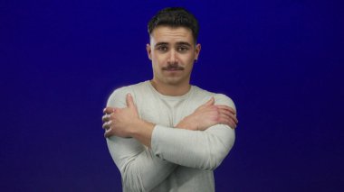 Genç İspanyol bir adam kollarını çapraz bir şekilde mavi bir arka plan duvarına yaslar, rahatlık ve iç gözlem yayar..