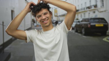 Yuvarlak metal çerçeveli gözlüklü genç adam, alnının üstündeki kıvırcık saçı bir şehir caddesinde park halindeki arabalara karşı ayarlar..