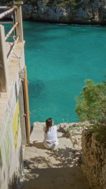 Santanyi Mallorca 'da, turkuaz su kenarında taş basamaklarda oturan kadın yazın huzurlu Akdeniz manzarasının tadını çıkarıyor..