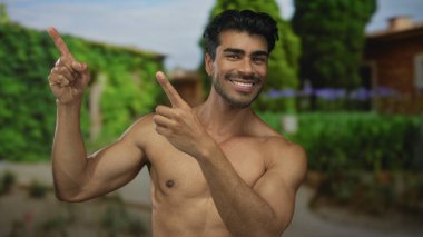 Tişörtsüz adam, yeşil yeşillik parkı dekoruyla kaplı binada geniş bir sırıtışla yukarı doğru işaret ediyor. Mutluluk..
