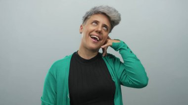 Yeşil hırka giyen bir kadın stüdyoda parmak kulağına yapılan telefon konuşmasını taklit ederken gülüyor..