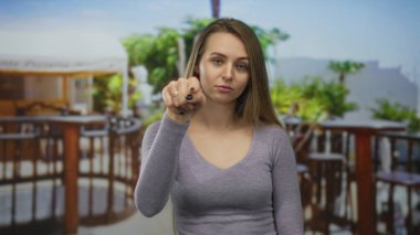 Mor bluzlu beyaz kadın, restoranın terasında durup kamerayı işaret ederken düşünüyor. Açık hava kafe manzarası bulanık ve arka planda yemyeşil bir alan var..
