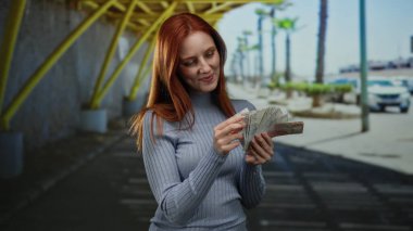 Elinde İsveç banknotları tutan bir kadın, şehir sokaklarında gülümsüyor, kızıl saçlı bir yetişkinin neşeli ifadesini sergiliyor..