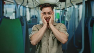 Otobüsün içinde sakallı genç bir adam şaşkınlığını ifade ediyor, elleri yüzün üzerinde, kapalı bir ulaşım ortamında, duygusal bir tepki sergiliyor..