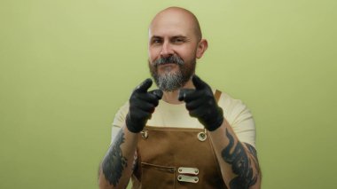 Kel, sakallı, eldivenli, yeşil bir duvara bakan, kahverengi önlüklü, elinde dövmeleri olan bir adam.