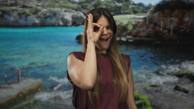 Genç İspanyol kadın, güneşli bir sahilde arka planda turkuaz denizle dikilirken gözünün önünde hoş bir jest yapıyor neşeli yaz titreşimleri sergiliyor..