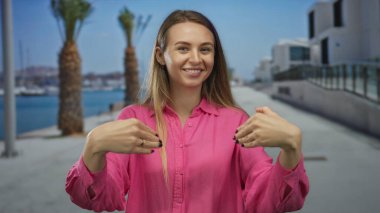 Pembe tişörtlü kadın, arka planda palmiye ağaçları ve suyla güneşli bir sahil yürüyüşünde gülümsüyor, deniz kenarında bir yerde neşe ve rahatlama yaşıyor..