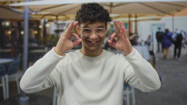 Gözlük takan İspanyol genç adam, iki elini de sokak ortasında şemsiyelerin altında işaret işareti yapıyor..