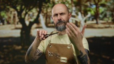 Sakallı, elinde makasla kel bir adam parkın dışında duruyor. Önlük takıyor, güneş ışığı altında kollarını gösteriyor. Arka planda bulanık ağaçlar var..