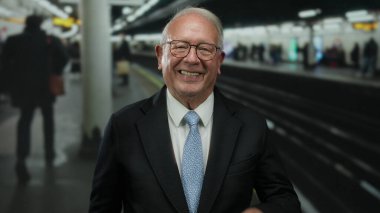 Takım elbiseli son sınıf öğrencisi başparmak kaldırıyor ve yoğun bir tren istasyonunu işaret ediyor. İçeride coşku ve pozitiflik gösteriyor..