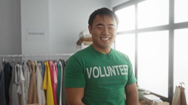 Bir hayır kurumunda gülümseyen genç bir adam yeşil gönüllü tişörtü giyip arka planda giyinmiş, toplum ruhunu ve hizmeti somutlaştırıyor..