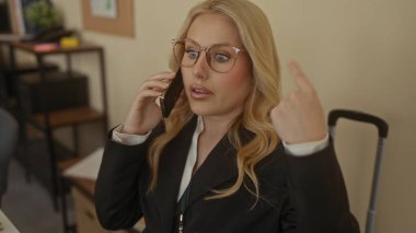 Ofisteki genç sarışın kadın yanında bavuluyla konuşuyor, gözlüklü ve resmi takım elbiseli, jestlerle şaşkınlığını dile getiriyor..