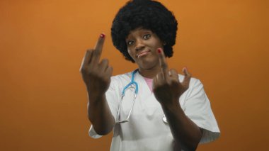 Beyaz önlük ve steteskop giyen genç Afro-Amerikan kadın doktor, turuncu stüdyoda orta parmağını kaldırıyor..
