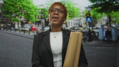 Siyah ceketli kadın, şehir caddesinde yeşil yeşilliklerin altında bina cephesi ve hırsla planları tutuyor..