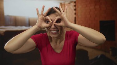 Kırmızı gömlekli bir kadın inşaat alanında gülümserken el dürbünü hareketi yapıyor; merak..