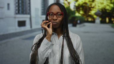 Şehir ortamında kırmızı gözlüklü, samimi bir gülümseme takan ve sessiz bir jest yapan kadın, örgü örüyor ve sokak ortasında günlük tarzını belirtiyor.