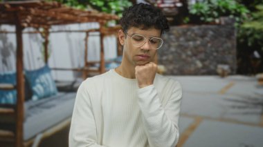 Beyaz süveterli ve gözlüklü İspanyol bir adam ahşap şezlongların yanında el pençe dinlenme; iç gözlem.