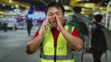 Yoğun bir havaalanı terminalinde, arka planı bulanık, sarı yelekli bir adam akıllı telefondan modern seyahat ortamı ve iletişim sahnesini gözler önüne seriyor..