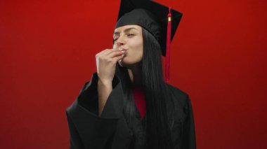 İspanyol kadın mezuniyetini parlak kırmızı bir arka planda kep ve cübbeyle kutluyor neşeli bir ifadeyle ve elini kaldırıyor..