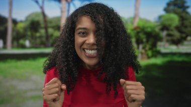 Kadın, kamera karşısında kıvırcık saçlı ve güneş ışığı altında kırmızı bluzlu bir şekilde gülümserken ormanda yumruklarını kaldırır..