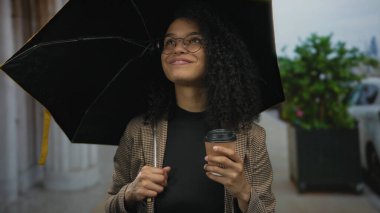 Sokakta yağmurlu bir günde elinde şemsiye ve kahve bardaklarıyla gülümseyen bir kadın..