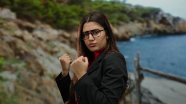 Sahilde güven içinde poz veren, gözlük takan, manzaralı bir okyanus zeminine karşı güç ve kararlılık sergileyen, zarafeti doğanın güzelliğiyle harmanlayan bir kadın..