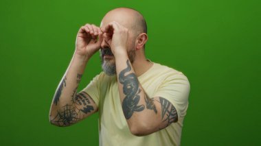 Dövmeli ve sakallı kel bir adam canlı yeşil bir arka planda hayali dürbünlere bakarak neşeli ve izole bir atmosfer yaratıyor..